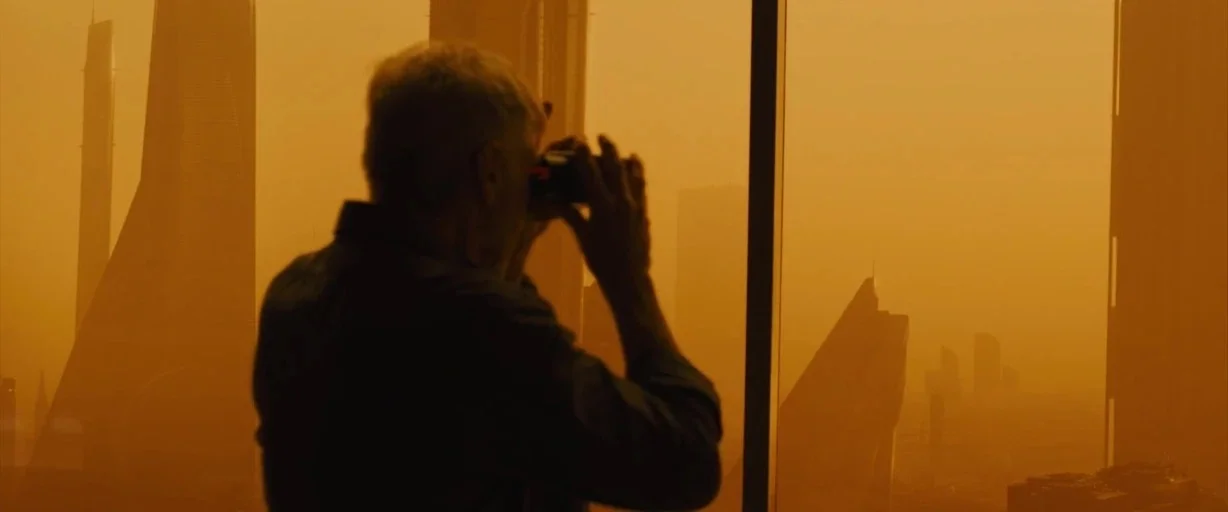 Blade Runner 2049 (2017) movie still: medium — A high-rise room overlooks a dust-filled cityscape under an orange-tinted sky. An elderly man stands…