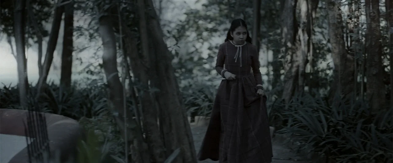 Evil Eye (2020) movie still: fullbody — A dense woodland path features overgrown foliage and thick dark tree trunks. A young girl in a period dress…