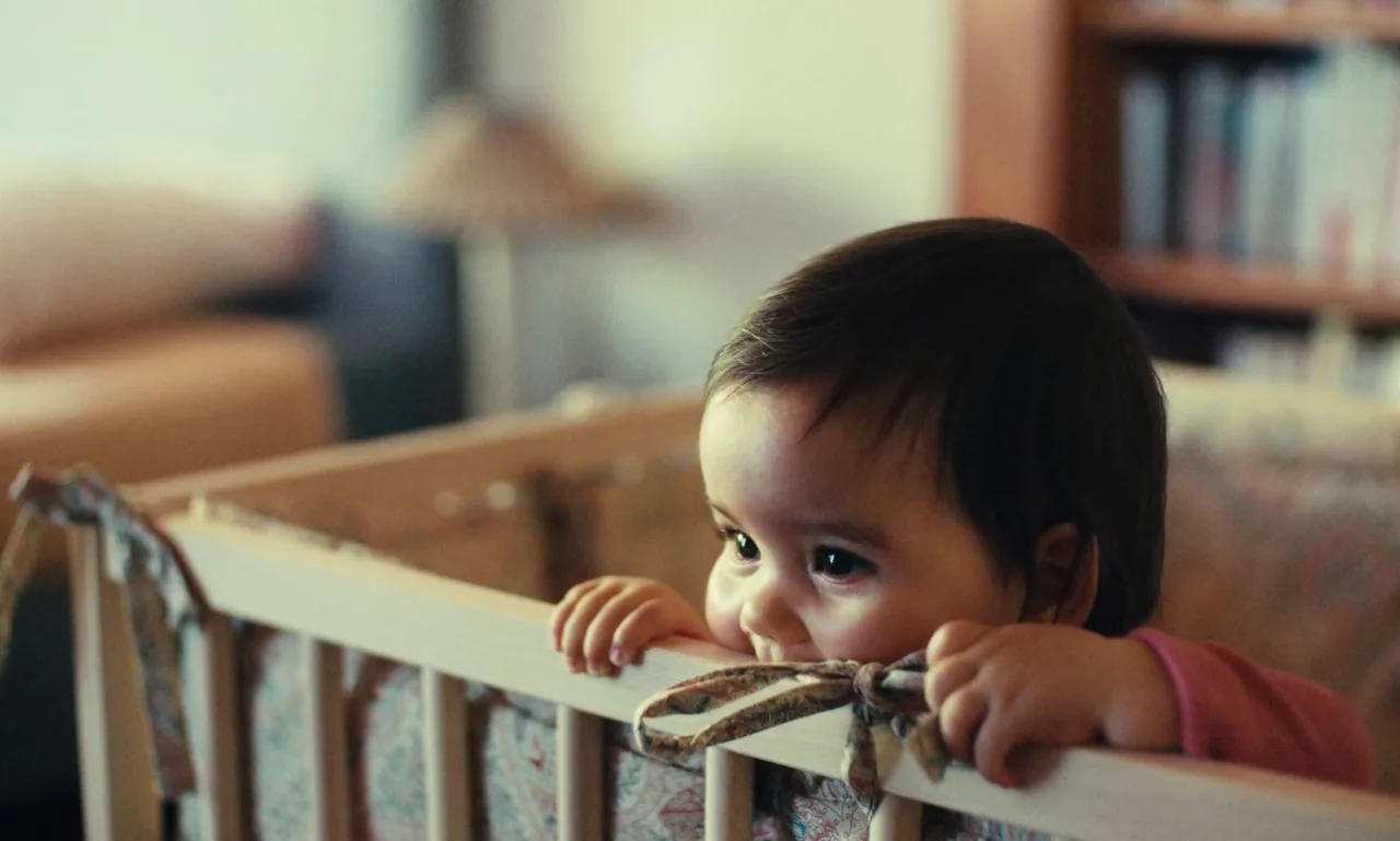 Just The Two Of Us (2023) movie still: closeup — A wooden baby crib sits inside a residential nursery space with blurred shelving behind. The child…