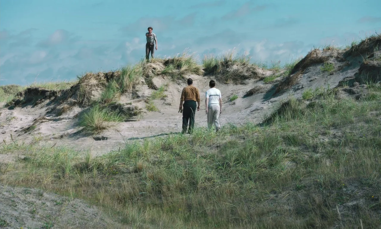 Bloodsuckers (2021) movie still: wide — A vast expanse of sandy coastal dunes covered in tufts of tall wild grass. Two men walk across the sand…