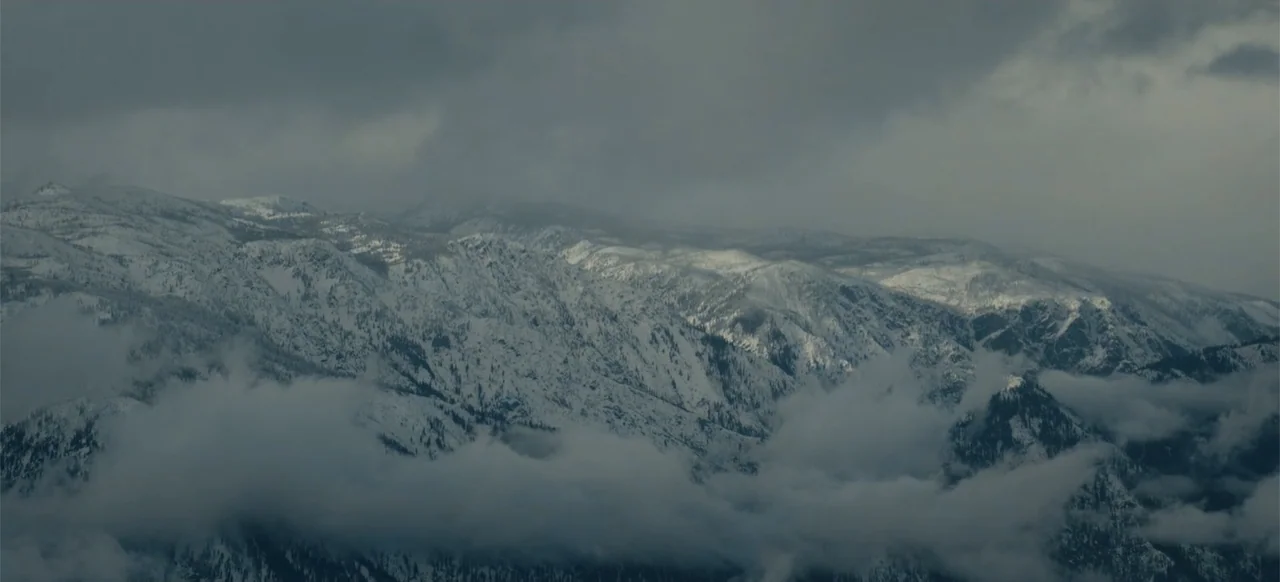 Devotion (2022) movie still: establishing — A vast mountain range covered in snow under a thick blanket of clouds. Dense fog rolls through the lower…