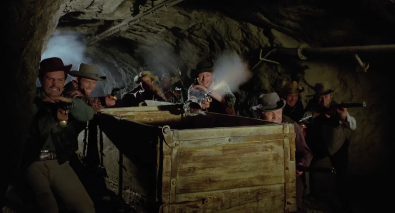 Johnny Colt (1966) movie still: medium — A group of armed men crouch behind a large wooden cart inside a dark cave. They are firing handguns toward…