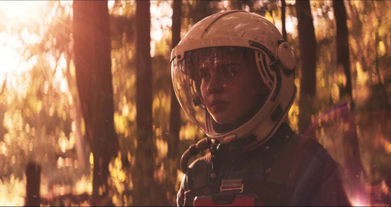 Prospect (2018) movie still: medium — A dense, sunlit woodland features tall vertical tree trunks and out-of-focus foliage. A young person wearing a…