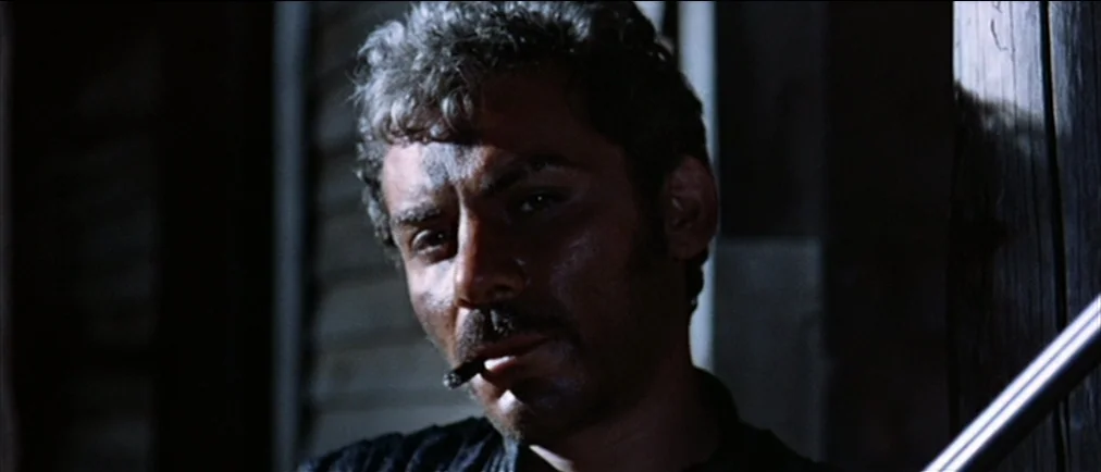 Fistful Of Dollars (1964) movie still: closeup — A dark wooden interior space with narrow gaps between the planks of the wall. A man with a mustache…