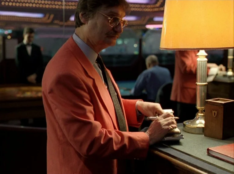 For Ever Mozart (1996) movie still: medium — An older man writes in a notebook at a dimly lit desk in a casino. Behind him, other people in…