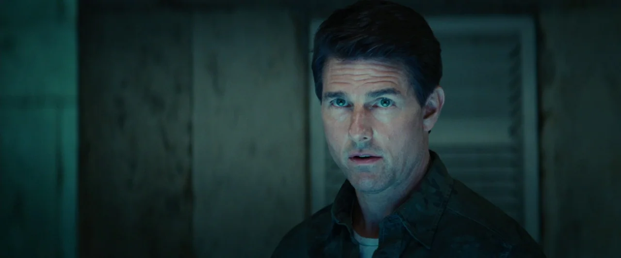 Edge of Tomorrow (2014) movie still: closeup — A dimly lit interior space with a muted, weathered wooden wall backdrop. A man looks toward the…