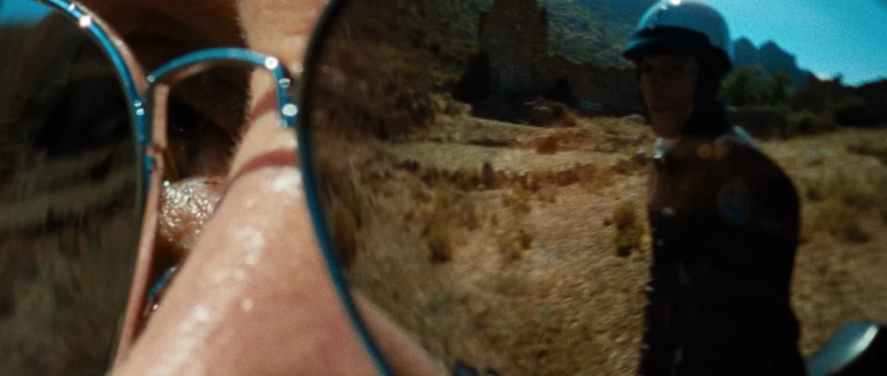 Let The Corpses Tan (2017) movie still: closeup — A dry, sun-baked landscape with sparse vegetation and rocky mountains in the background. A person…
