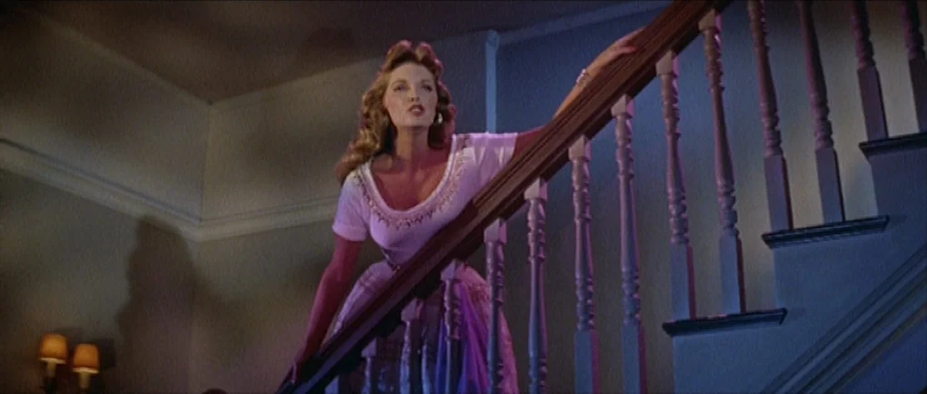 The Girl Can’t Help It (1956) movie still: medium — An interior staircase with wooden banisters and spindles leads up into shadows. A woman descends…