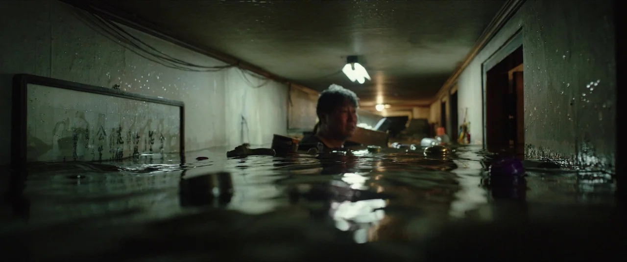 Parasite (2019) movie still: medium — A low-ceilinged apartment interior is completely submerged in dark, murky water. A man wades through the…