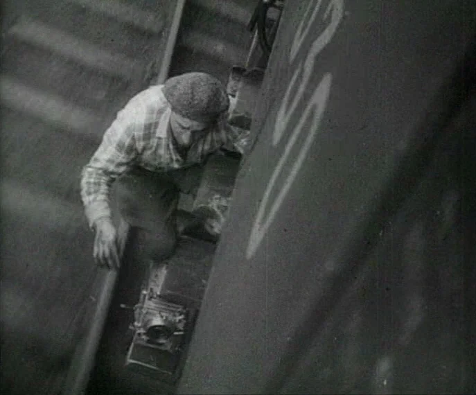 Man with a Movie Camera (1929) movie still: medium — The exterior of a train carriage with metallic surfaces and visible markings. A man crouches…