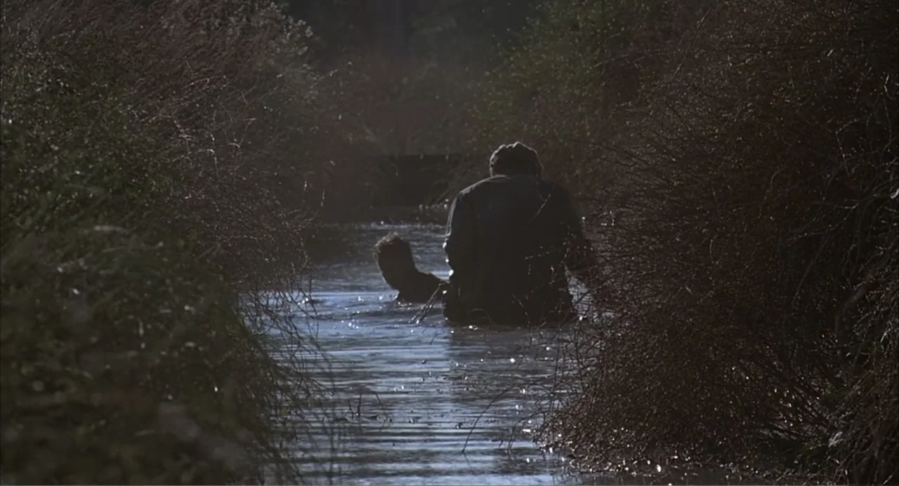 Of Mice and Men (1992) movie still: medium — A narrow creek flows between dense, dry brush and tall, dead-looking riparian vegetation. Two figures…