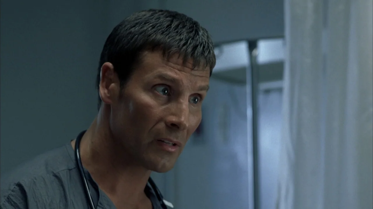 John Q (2002) movie still: closeup — A clinical hospital room with a white privacy curtain in the background. A male doctor in scrubs looks toward…
