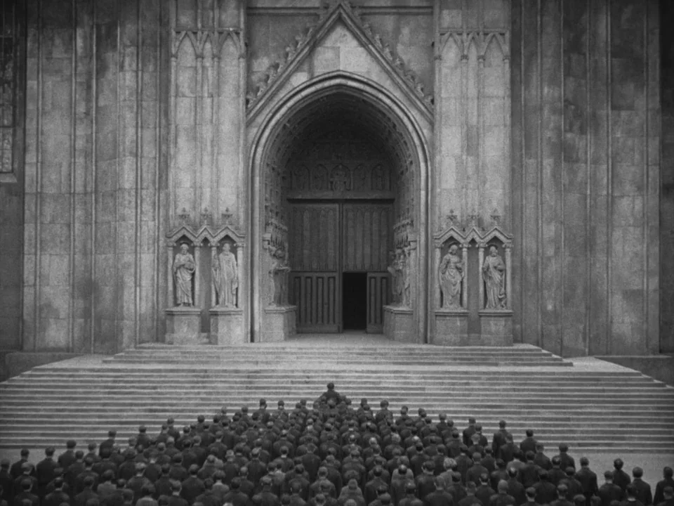 Metropolis (1927) movie still: wide — A massive Gothic cathedral entrance features ornate stone statues flanking large wooden doors. A large crowd…