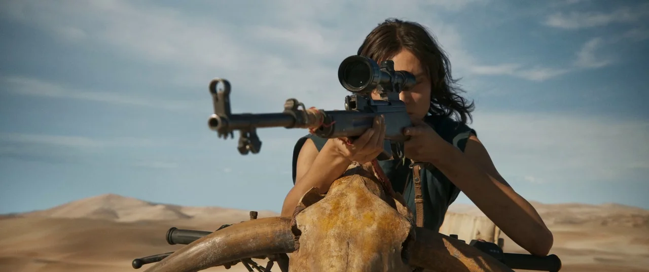 Furiosa: A Mad Max Saga (2024) movie still: medium — A vast, arid desert landscape under a clear, pale blue sky. A woman aims a rifle using a…