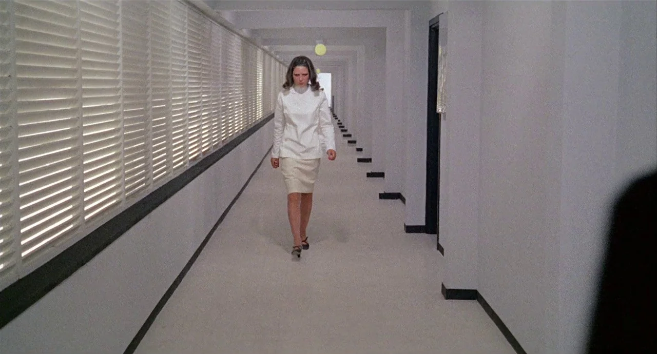 The 10th Victim (1965) movie still: fullbody — A long, sterile hospital corridor features rows of white slatted wall blinds and tiles. A woman in a…