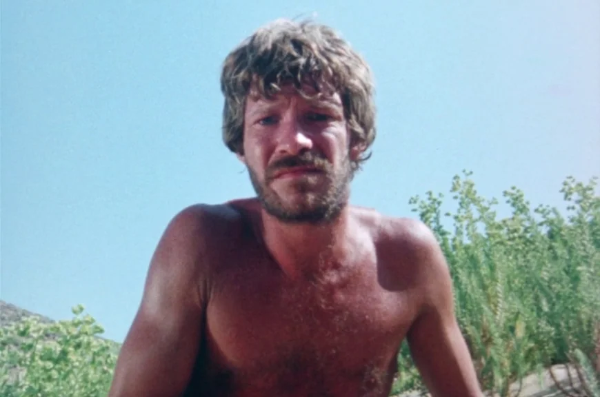 Sebastiane (1976) movie still: medium — A man stands outdoors with sparse green vegetation and a rocky slope behind him. The man is shirtless and…