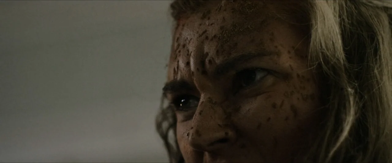 Ambush (2020) movie still: closeup — A close up view of a woman's face covered in dark soil or grit. She looks directly ahead with a focused and…