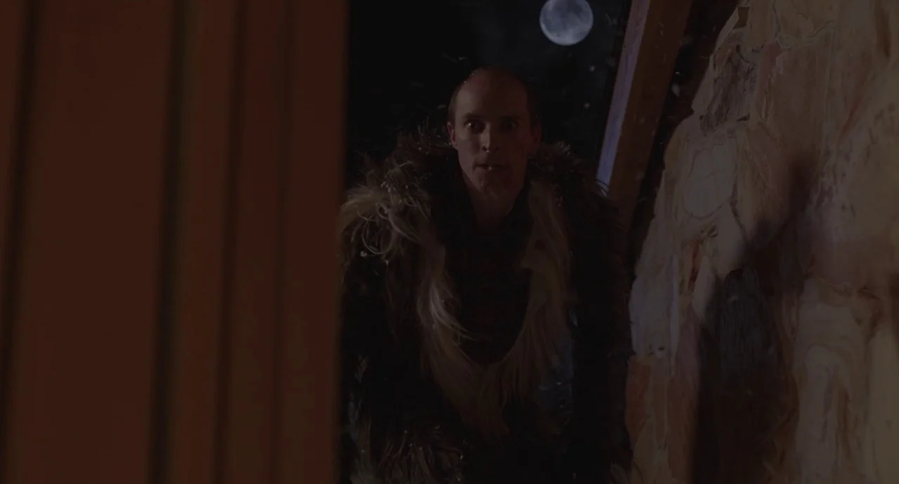 The Wolf of Snow Hollow (2020) movie still: medium — A man stands before a rough stone wall under a dark night sky. The moon is visible in the upper…