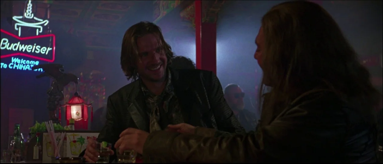 Strange Days (1995) movie still: medium — A dimly lit, smoky bar features a neon Budweiser sign on the wall. Two men sit at the counter conversing…