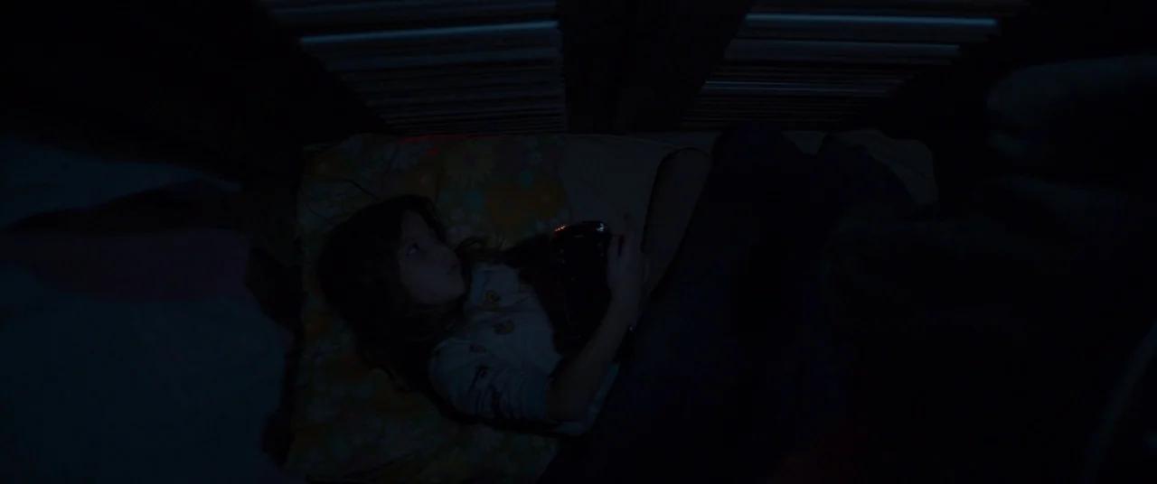 Room (2015) movie still: medium — A young girl lies in a dark bed beneath horizontal wooden slats. She holds a small glowing object that illuminates…