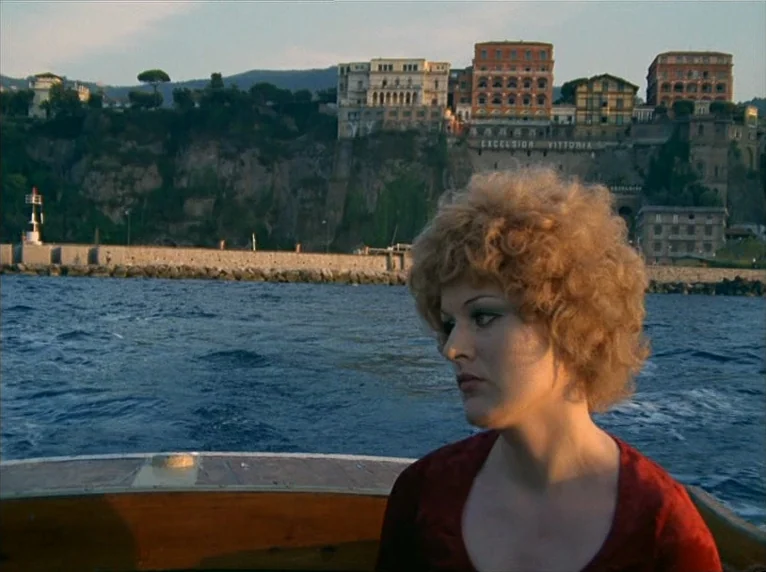 Beware Of A Holy Whore (1971) movie still: medium — A woman sits in a wooden boat on open water during sunset. Coastal buildings on a rocky cliff…