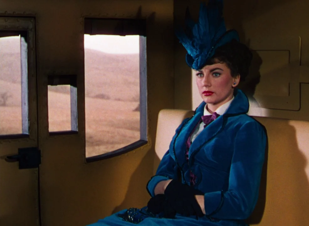 Calamity Jane (1953) movie still: medium — A woman sits inside the compact cabin of a horse-drawn coach. Through the windows, a dry landscape passes…