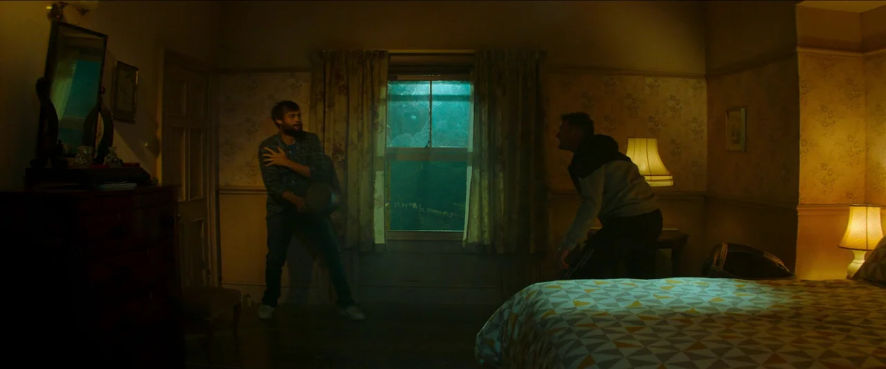 Unwelcome (2022) movie still: wide — A dimly lit room features patterned wallpaper and a window covered by floral curtains. Two men stand facing…