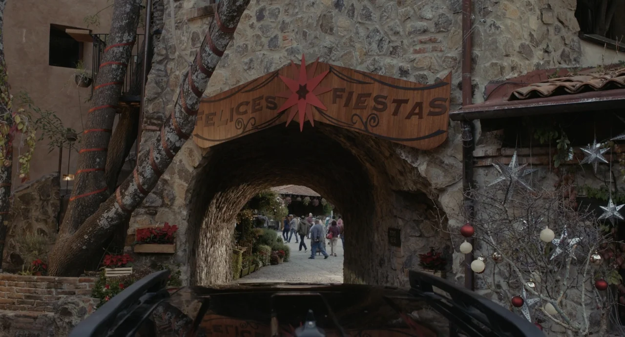 Dating Santa (2023) movie still: establishing — A stone masonry archway features a wooden sign reading Felices Fiestas above the passage.…