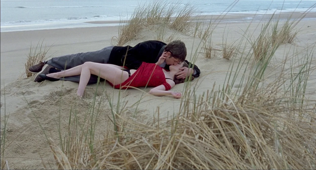 Fat Girl (2001) movie still: fullbody — A beach with sand dunes covered in tall, dry beach grass near the ocean. A young couple lies on their sides…