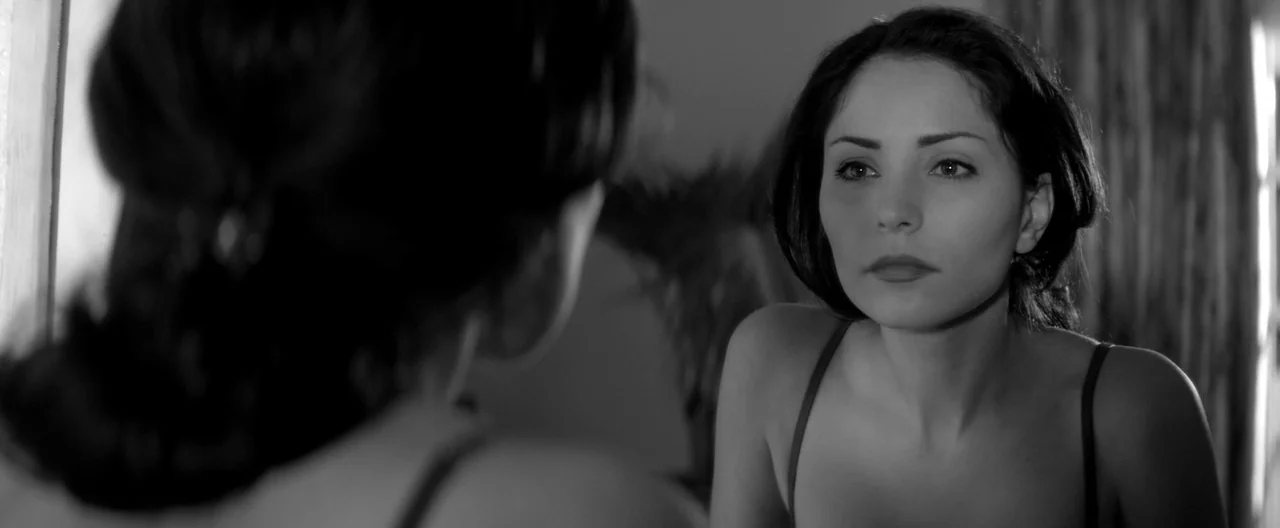 A Girl Walks Home Alone at Night (2014) movie still: medium — A woman stares at her own reflection in a mirror in a dimly lit room. She wears a thin…