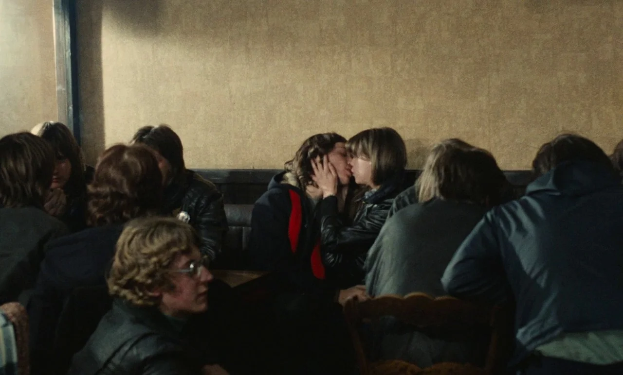 Graduate First (1978) movie still: medium — A dimly lit, narrow indoor space features people gathered around a dark wooden table. A couple kisses in…