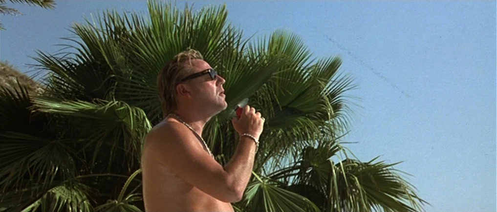Sexy Beast (2000) movie still: medium — A man stands outdoors in front of a large, dense green palm tree. The man holds a small red object toward…