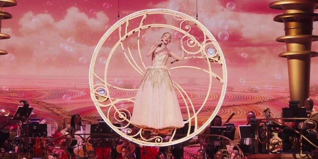 Wicked: One Wonderful Night (2025) movie still: fullbody — A theatrical stage with a pink painted backdrop featuring stylized rolling hills and…