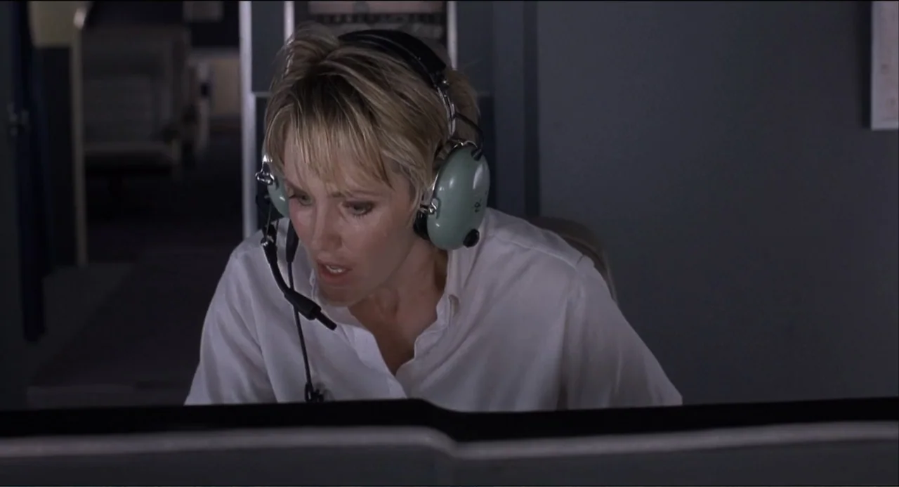 Air Rage (2001) movie still: medium — A dimly lit industrial or office space with grey panels in the background. A woman in a white shirt wears a…