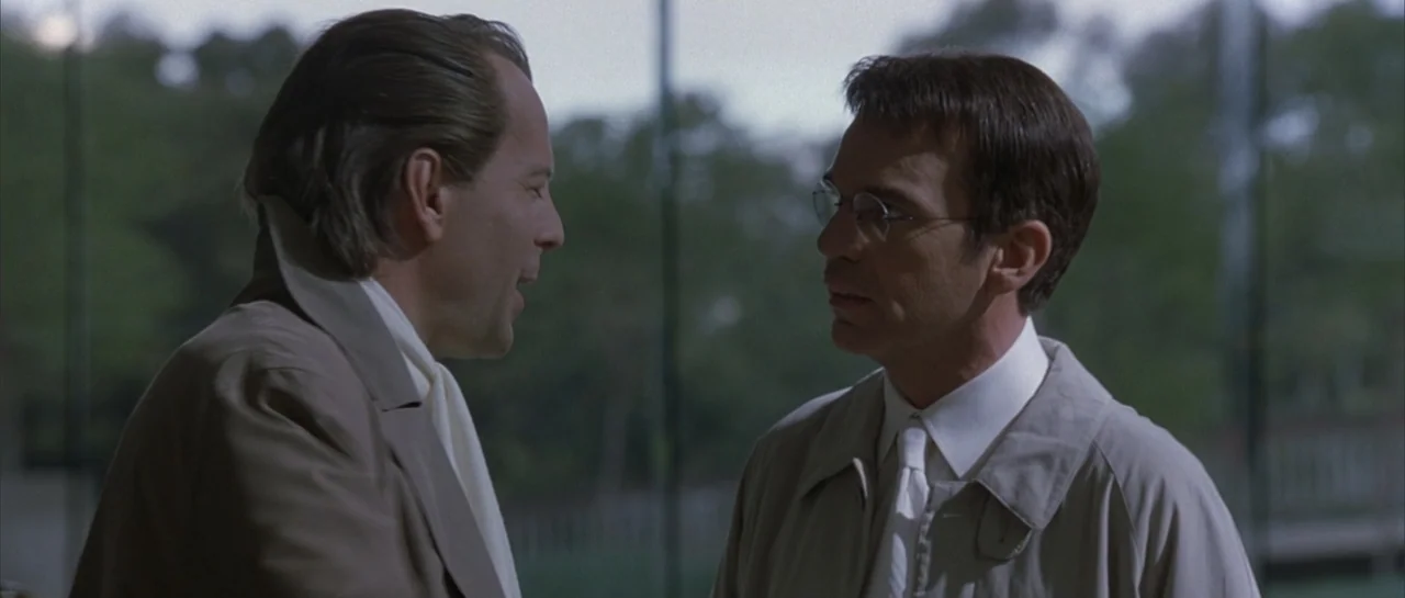 Bandits (2001) movie still: medium — An outdoor park setting with blurred green trees in the background. Two men in matching tan trench coats stand…