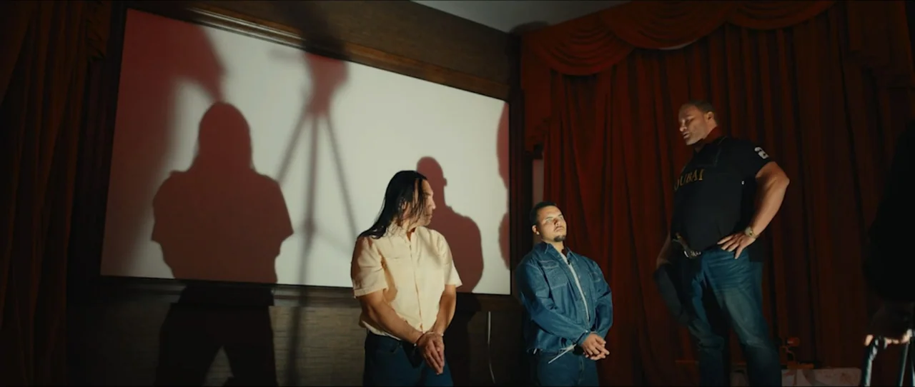 American Sicario (2021) movie still: medium — A dimly lit room features a large projection screen flanked by heavy red curtains. Two men stand…