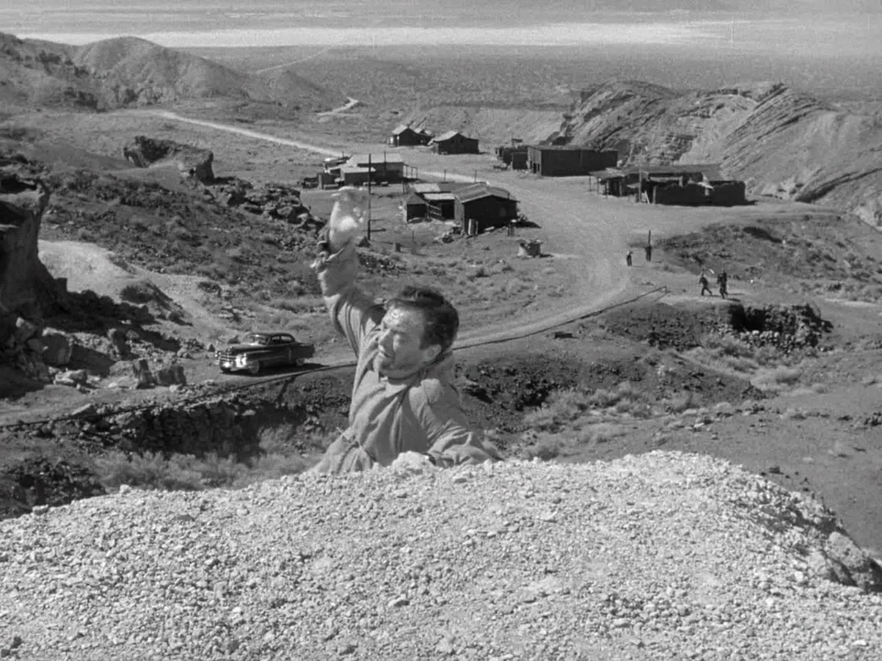 The Prowler (1951) movie still: wide — A dusty desert hillside overlooks a cluster of small wooden shacks and a road. A man in a trench coat climbs…