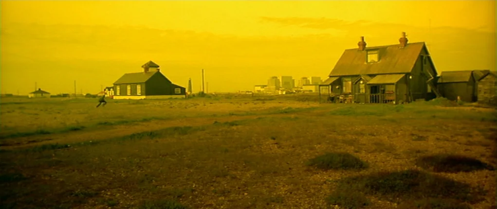I Want You (1998) movie still: wide — A desolate flat coastal area features a few scattered wooden structures and buildings. A lone figure is…