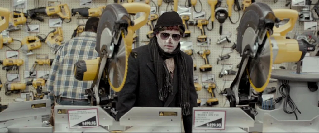 Clown (2014) movie still: medium — A well-lit aisle in a store filled with yellow power tools on pegboards. A man in a dark coat and hat stands…