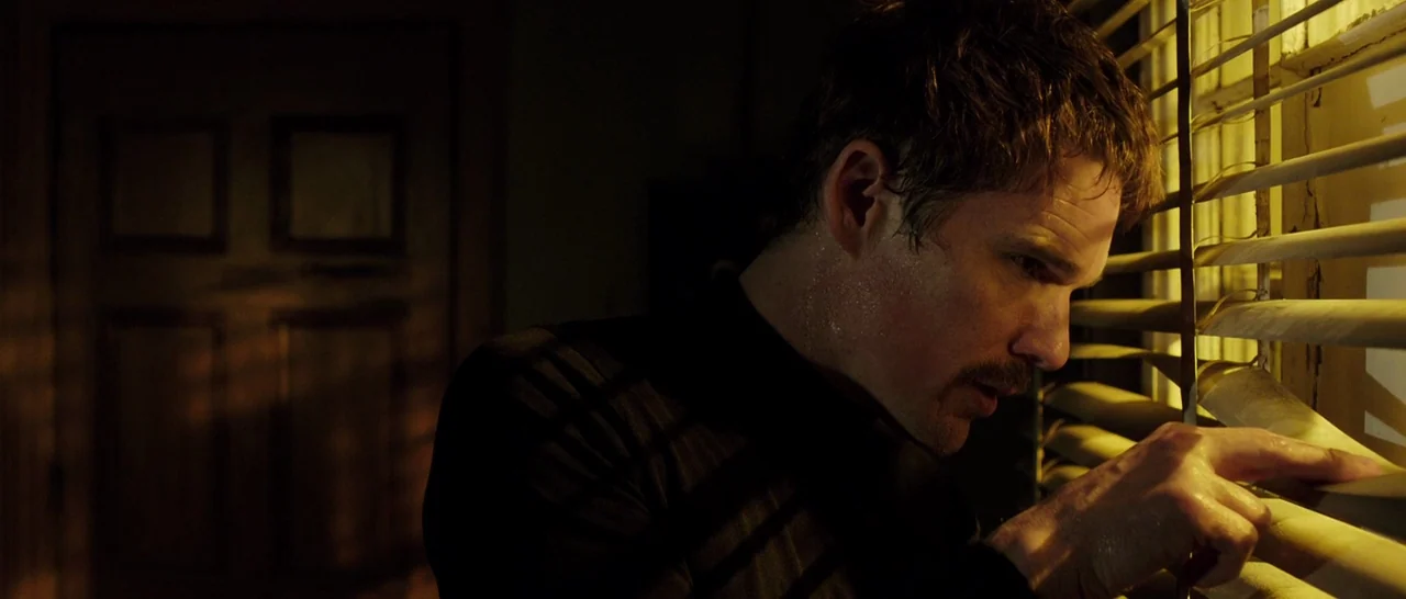 Predestination (2014) movie still: medium — A dimly lit room at night features a man standing by a window. He uses his fingers to pull back the…