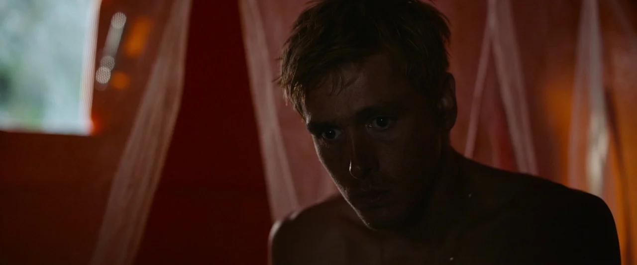 Triangle of Sadness (2022) movie still: closeup — The interior of a translucent orange fabric shelter filters external light. A shirtless young man…