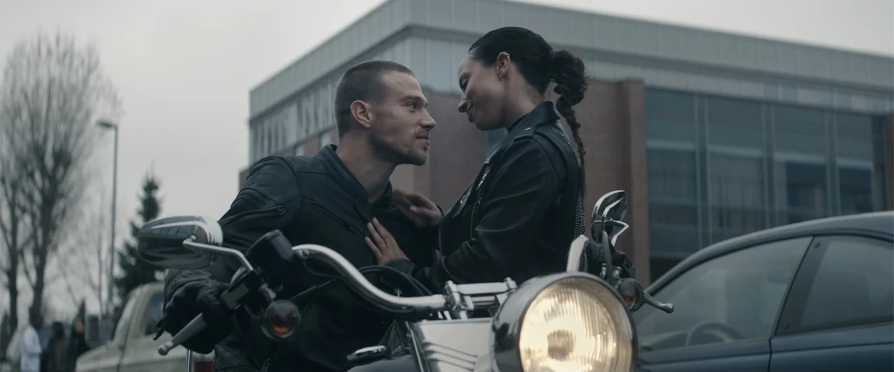 Perfect Addiction (2023) movie still: medium — An outdoor parking area features a modern multi-story brick building in the background. A man and a…