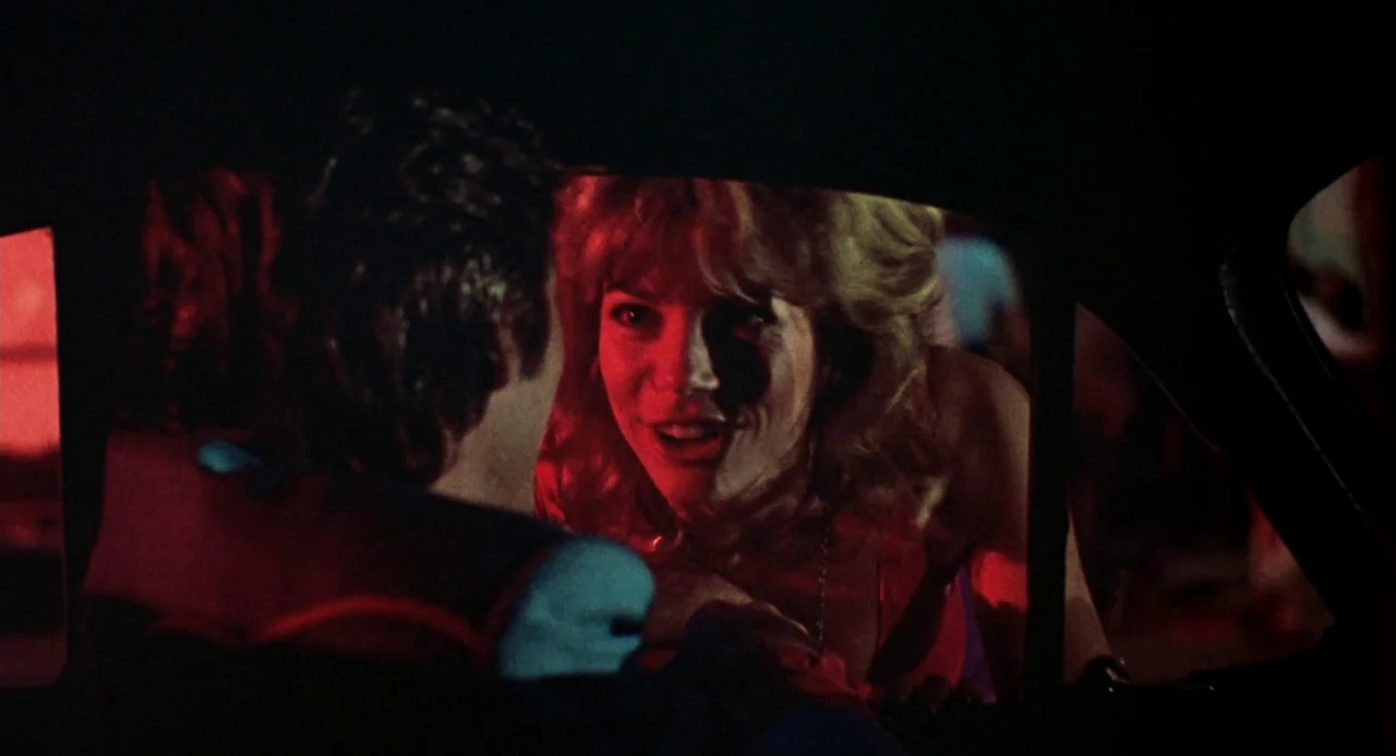 Breathless (1983) movie still: medium — A woman leans through a car window to look at a male driver at night. Deep red neon light illuminates the…