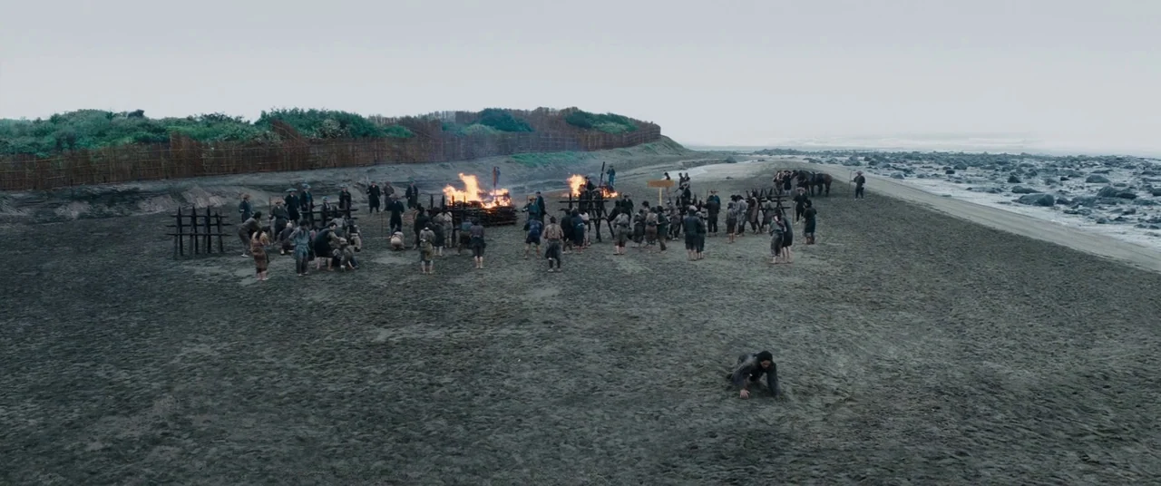 Silence (2016) movie still: wide — A dark, expansive beach features a crowd of people gathered around several small fires. A lone person crawls…