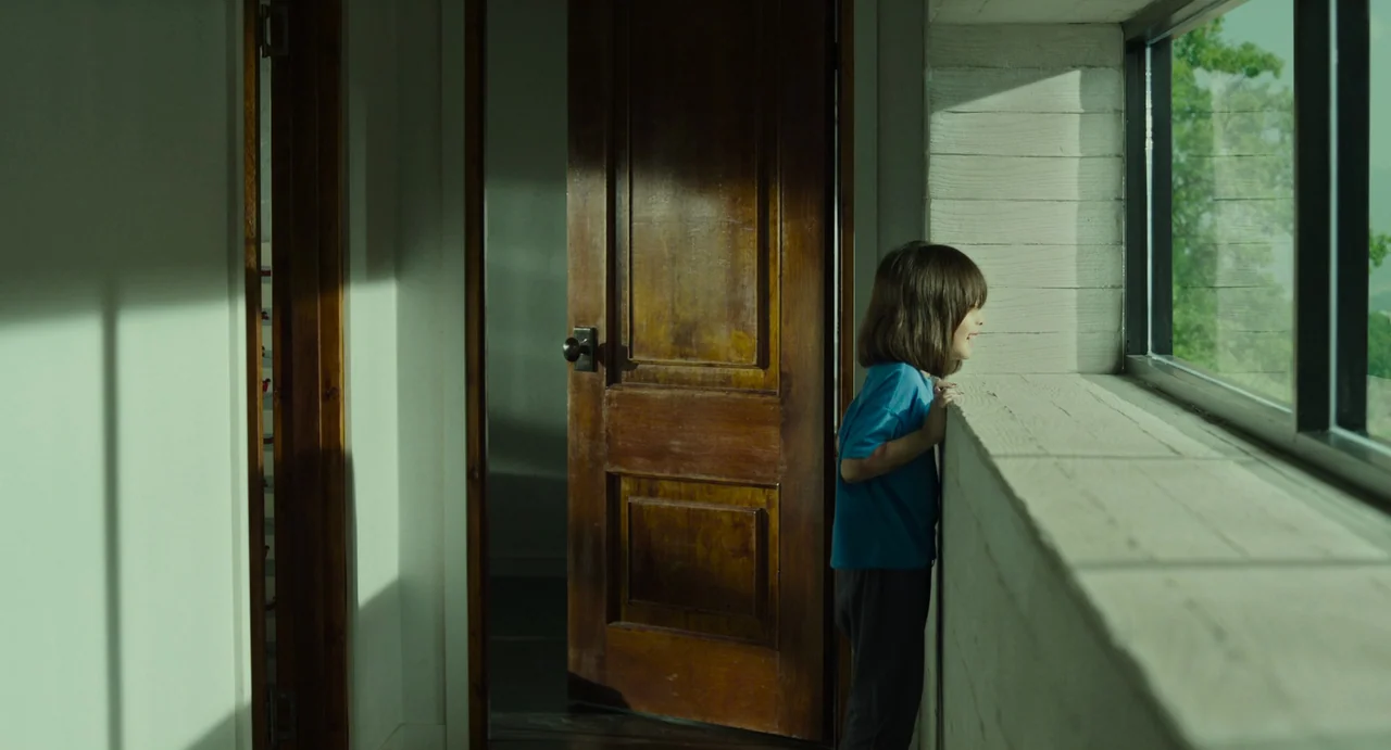 The Mimic (2017) movie still: medium — A quiet indoor corridor with white walls and a polished wooden door frame. A small child stands by a window…