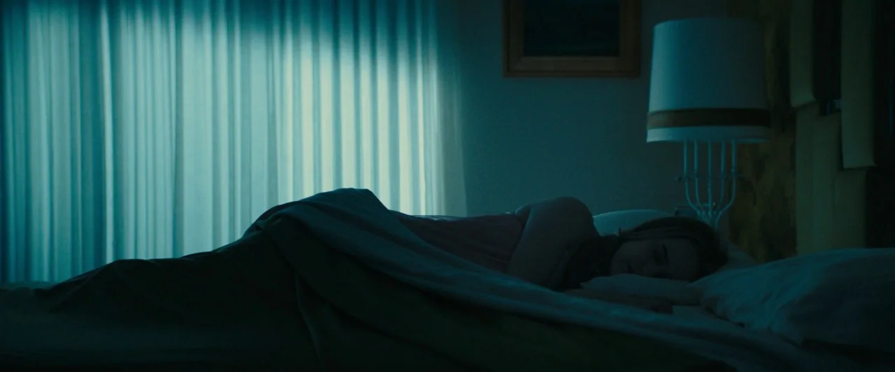 I'm Your Woman (2020) movie still: medium — A dark hotel room is illuminated by a faint glow through floor-to-ceiling curtains. A woman lies asleep…