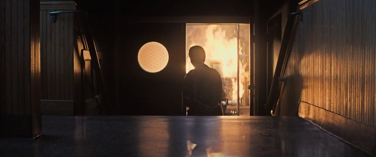 The Many Saints of Newark (2021) movie still: fullbody — A dimly lit indoor stairwell with wood-paneled walls and a circular light fixture. A man in…