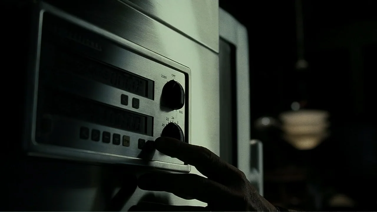 Mr. Brooks (2007) movie still: closeup — A metallic control panel with digital displays and black knobs fills the frame. A human finger presses a…