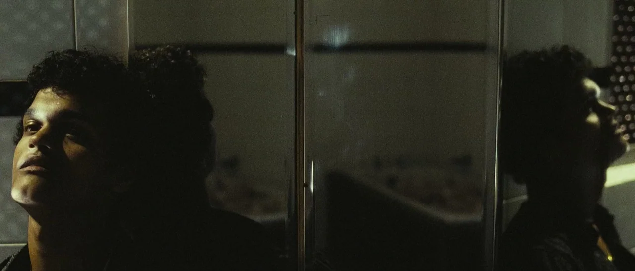 Slumdog Millionaire (2008) movie still: closeup — A dark, tiled bathroom features a reflective surface split vertically by a frame. A young man…