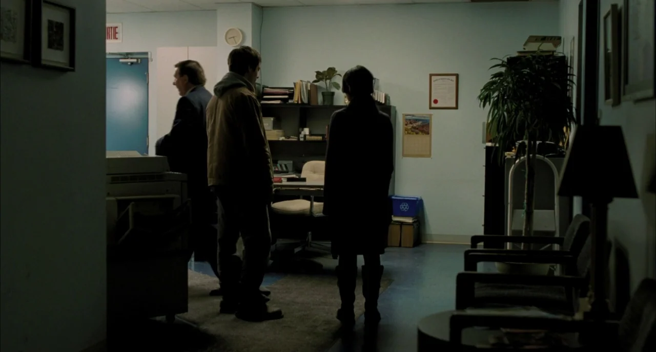 Incendies movie still: medium — An office space contains a desk, chairs, and a potted plant against light walls. Three individuals stand in the room…